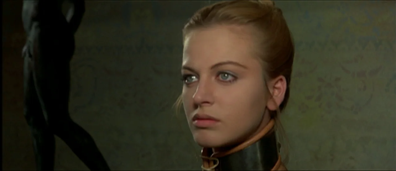 Flesh For Frankenstein (1973) movie still: closeup — A woman stands in a gallery space with patterned walls and a dark statue. She looks toward the…