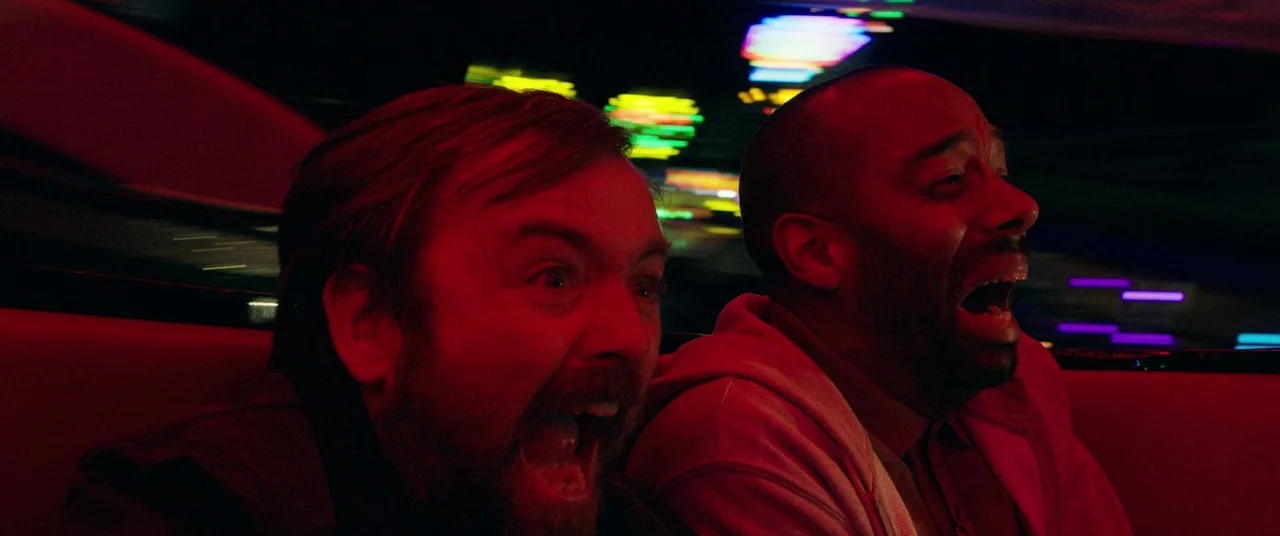 Bull (2021) movie still: medium — Two men sit side-by-side inside a vehicle driving at high speed at night. Bright blurry neon light streaks reflect…