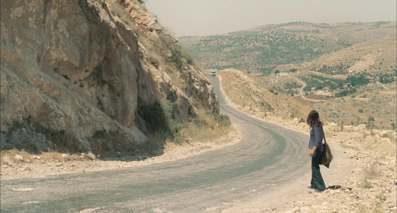 Incendies movie still: wide — A winding asphalt road cuts through a dry, hilly, and arid terrain landscape. A woman stands alone on the gravelly…