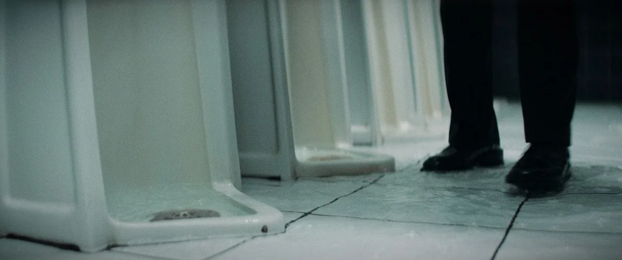 La Llorona (2019) movie still: medium — A tiled room contains a line of white porcelain floor-mounted urinals. A person wearing dark trousers and…