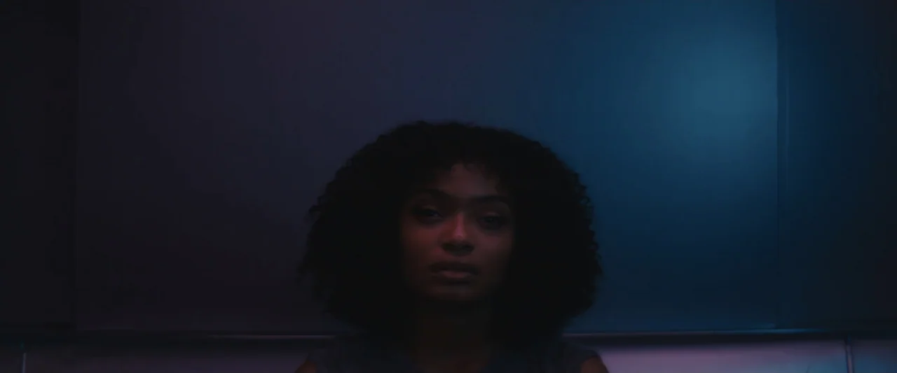 The Sun Is Also a Star (2019) movie still: medium — A dark, minimally furnished interior space features a solid wall background. A woman with…