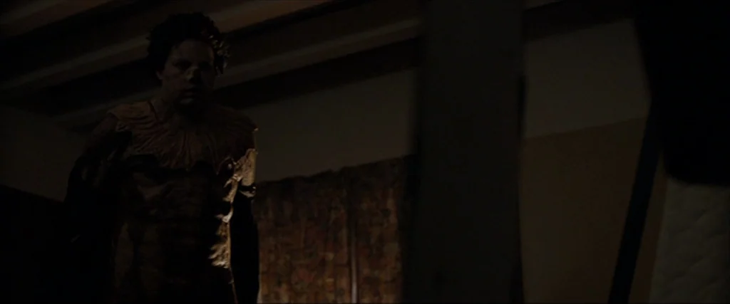 Clown (2014) movie still: medium — A man in a textured, frilled-collar garment stands in a dimly lit dark room. He looks toward the camera while the…