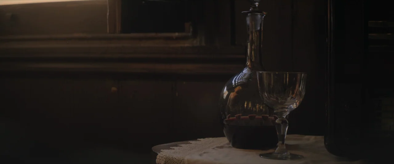 The New Boy (2023) movie still: closeup — A glass decanter filled with red wine and a stemmed glass rest on a tabletop. The scene is illuminated by…