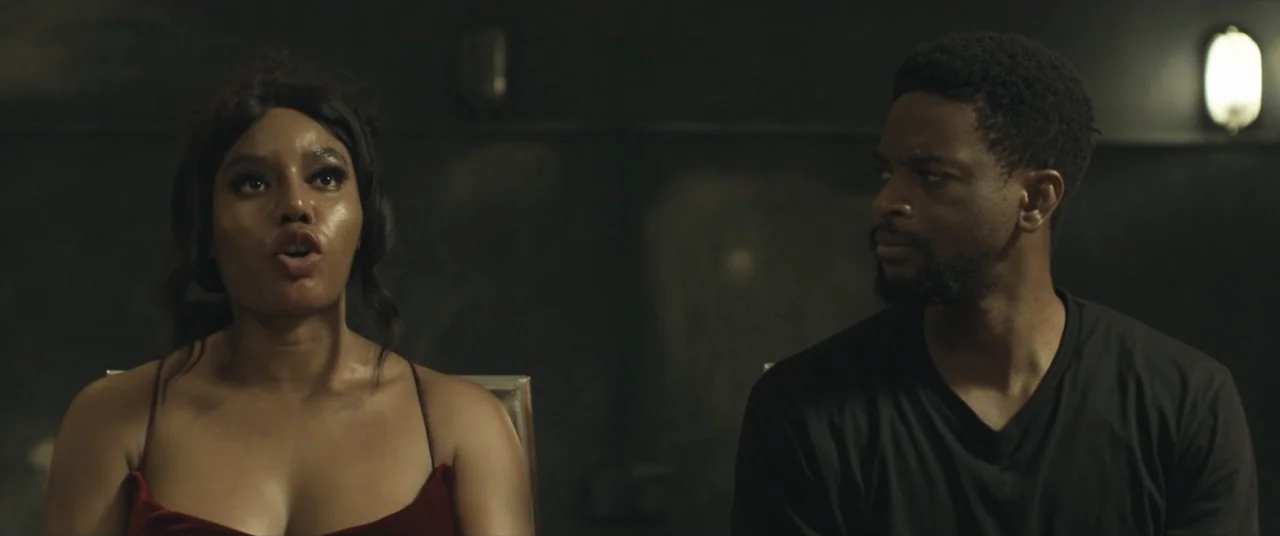 Saving Onome (2024) movie still: medium — A dark, sparsely lit interior space with a black background wall. A woman and a man sit side by side…