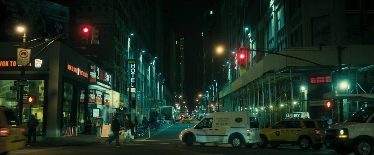 Whiplash (2014) movie still: establishing — A night view of a dense urban intersection lined with neon-lit commercial businesses. Yellow taxis and a…