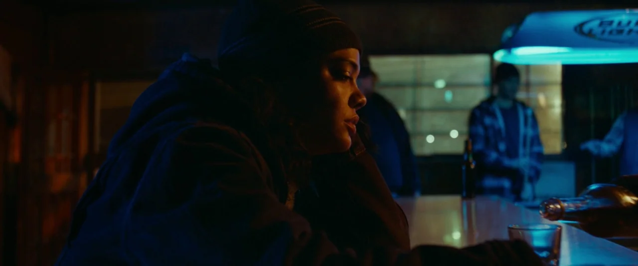 Little Woods (2018) movie still: medium — A dimly lit counter with a blue neon sign glowing in the background. A person sits at the counter resting…