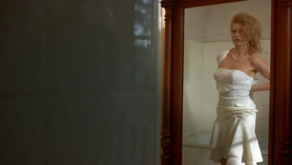 The Marriage of Maria Braun (1979) movie still: medium — A woman in a white dress stands before a tall wood-framed mirror in a tiled room. She fixes…