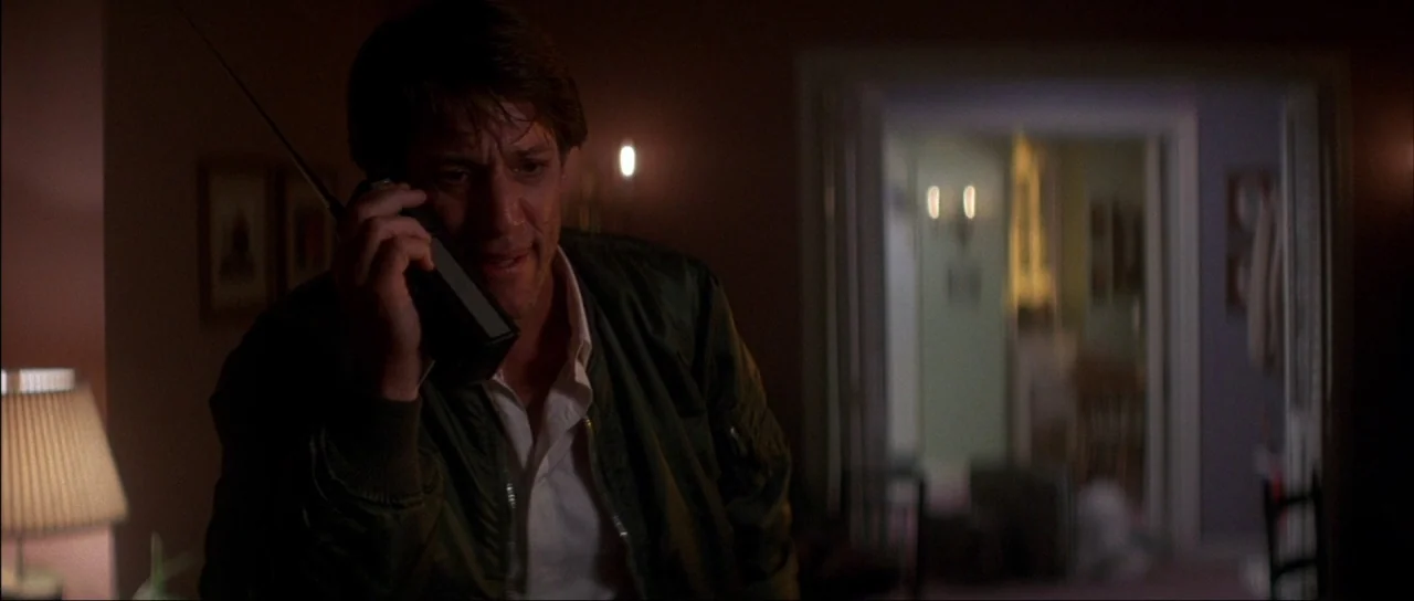 The Monster Squad (1987) movie still: medium — A dimly lit room contains a man holding an old-fashioned walkie-talkie. In the background a doorway…