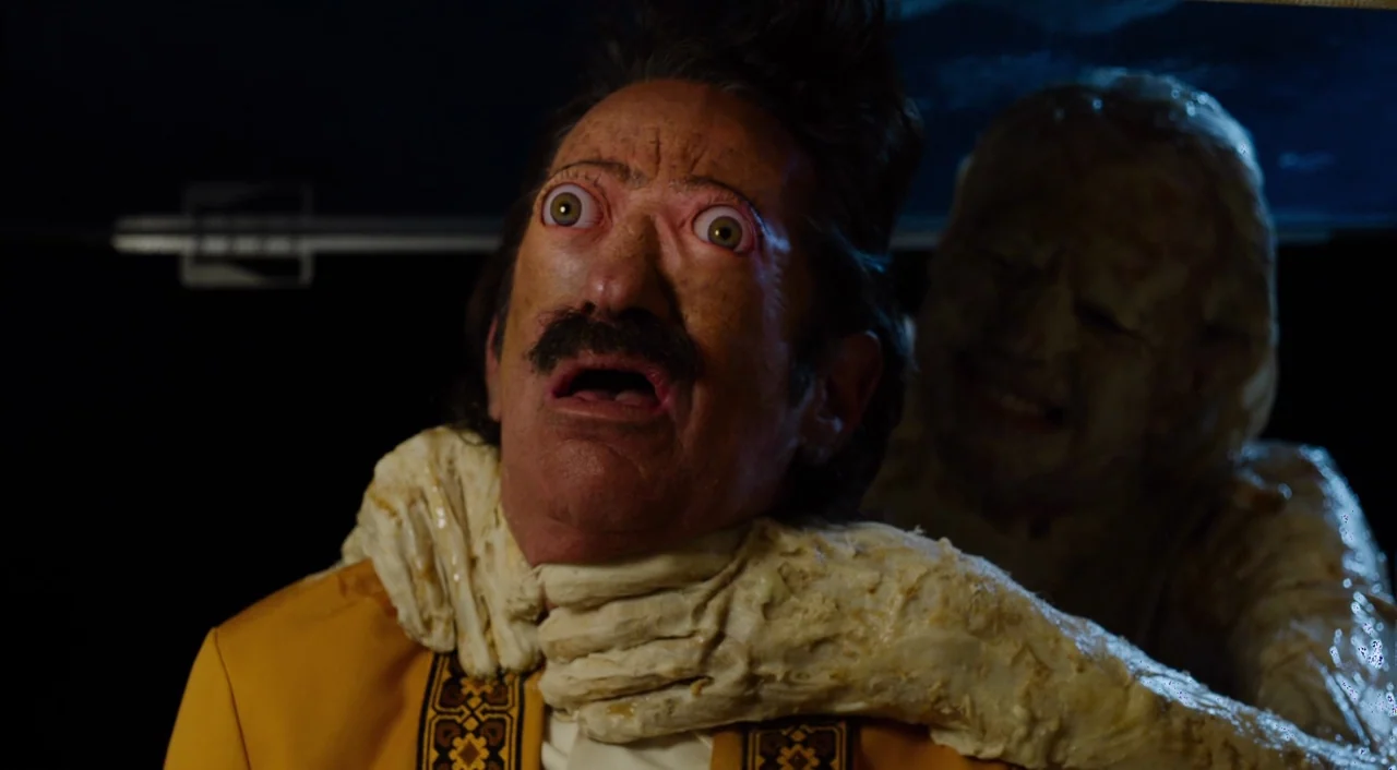 The Greasy Strangler (2016) movie still: medium — A man in a yellow blazer sits in a dark vehicle cabin. A pale, textured entity reaches around from…