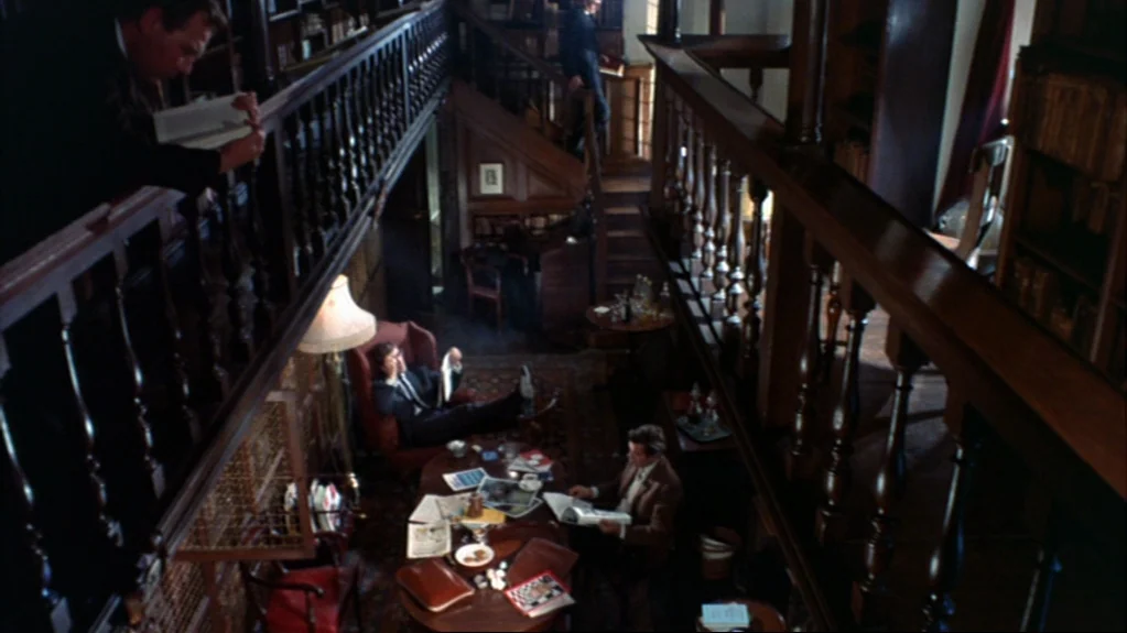 Accident (1967) movie still: wide — A multi-level wood-paneled library features a central lounge area with armchairs and tables. Three men occupy…