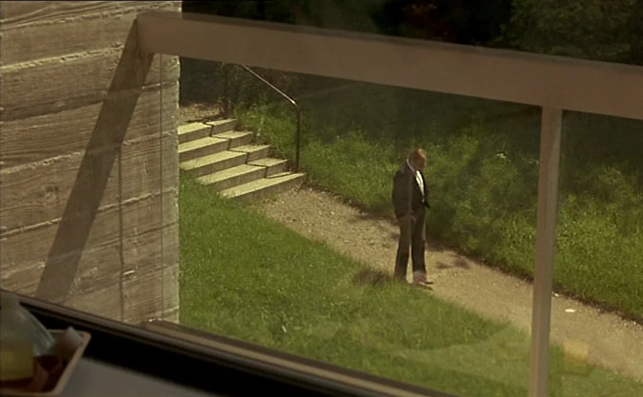 The Woman Next Door (1981) movie still: wide — A view from an interior balcony overlooking a grassy yard with concrete stairs. A middle-aged man in…