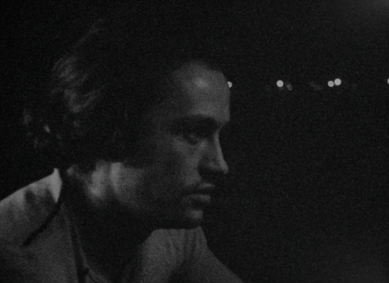 Je Tu Il Elle (1974) movie still: closeup — A man stands in darkness against a blurry background of distant city night lights. He faces right while…