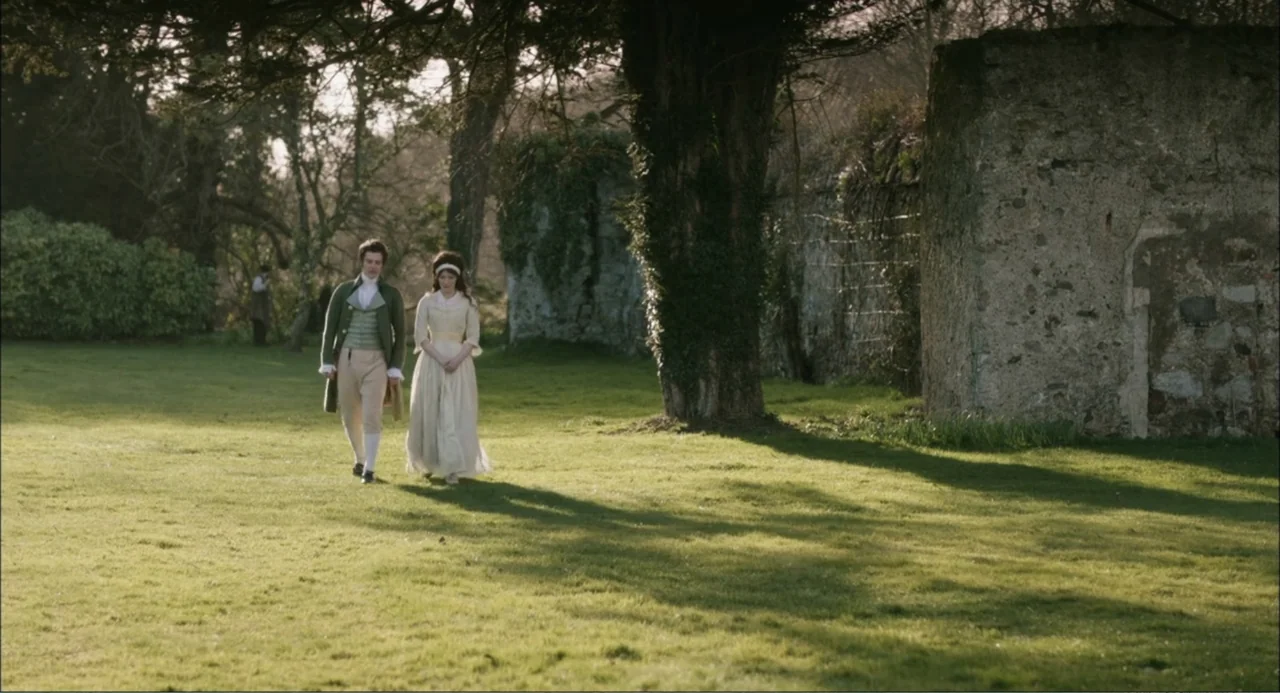 Love & Friendship (2016) movie still: fullbody — A green open park space features large trees and a crumbling stone wall background. A man in period…