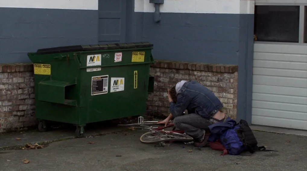 Your Sister’s Sister (2011) movie still: fullbody — A person crouches beside a large green dumpster positioned against a brick and concrete wall.…