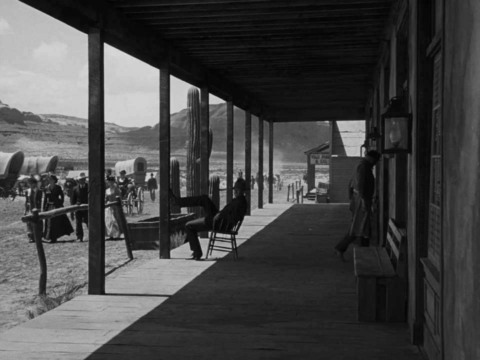 My Darling Clementine (1946) movie still: wide — A wooden covered porch overlooks a dusty frontier town with covered wagons in distance. A man in a…
