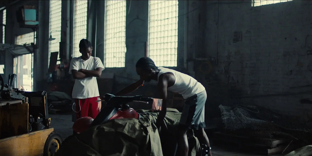 Charm City Kings (2020) movie still: fullbody — A spacious industrial warehouse features concrete block walls and grid-patterned glass windows. Two…