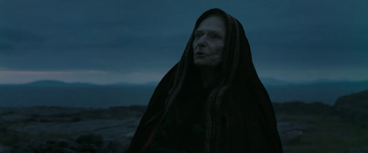 The Banshees of Inisherin (2022) movie still: medium — An elderly woman stands alone on a desolate, rocky landscape near the water. She stares…