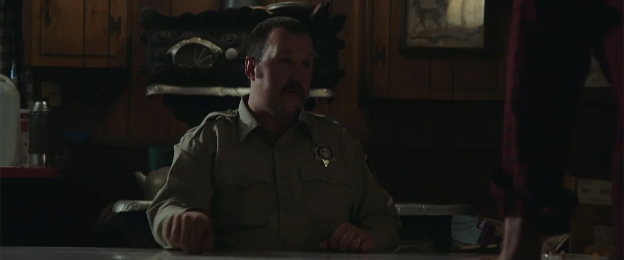 Darkness Waits (2020) movie still: medium — A dark, wood-paneled room serves as the setting for a quiet meeting. A male police officer in a uniform…