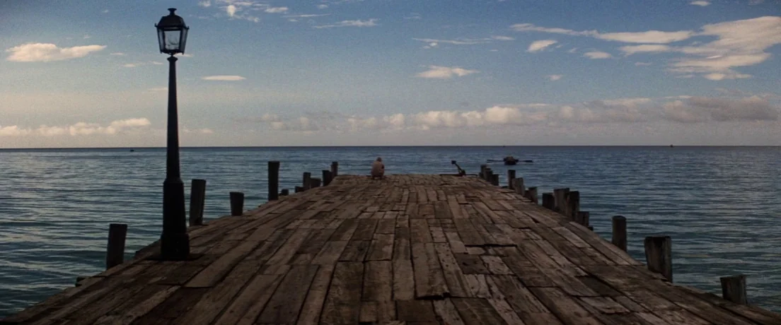 Papillon (1973) movie still: wide — A long wooden pier extends into a vast calm ocean under a clear sky. A lone figure squats at the far end of the…