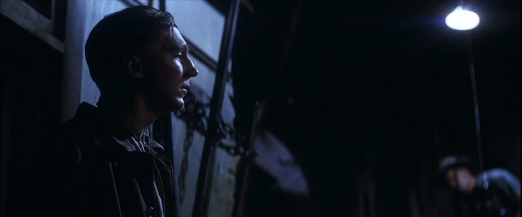 There Will Be Blood (2007) movie still: medium — A dimly lit loading dock at night with heavy iron chains hanging nearby. The foreground subject…