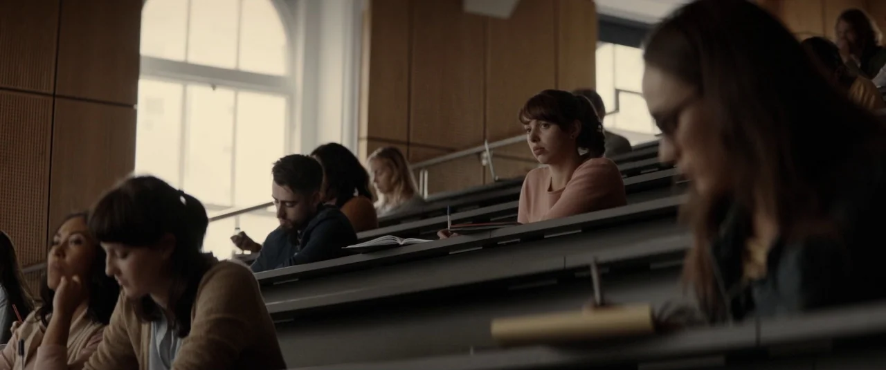 The Hole in the Ground (2019) movie still: medium — A modern university lecture hall with tiered seating and wooden wall paneling. Multiple students…