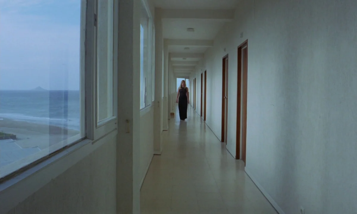 Vampyros Lesbos (1971) movie still: fullbody — A long, minimalist hotel corridor with white walls stretches toward a distant point. A woman in a…