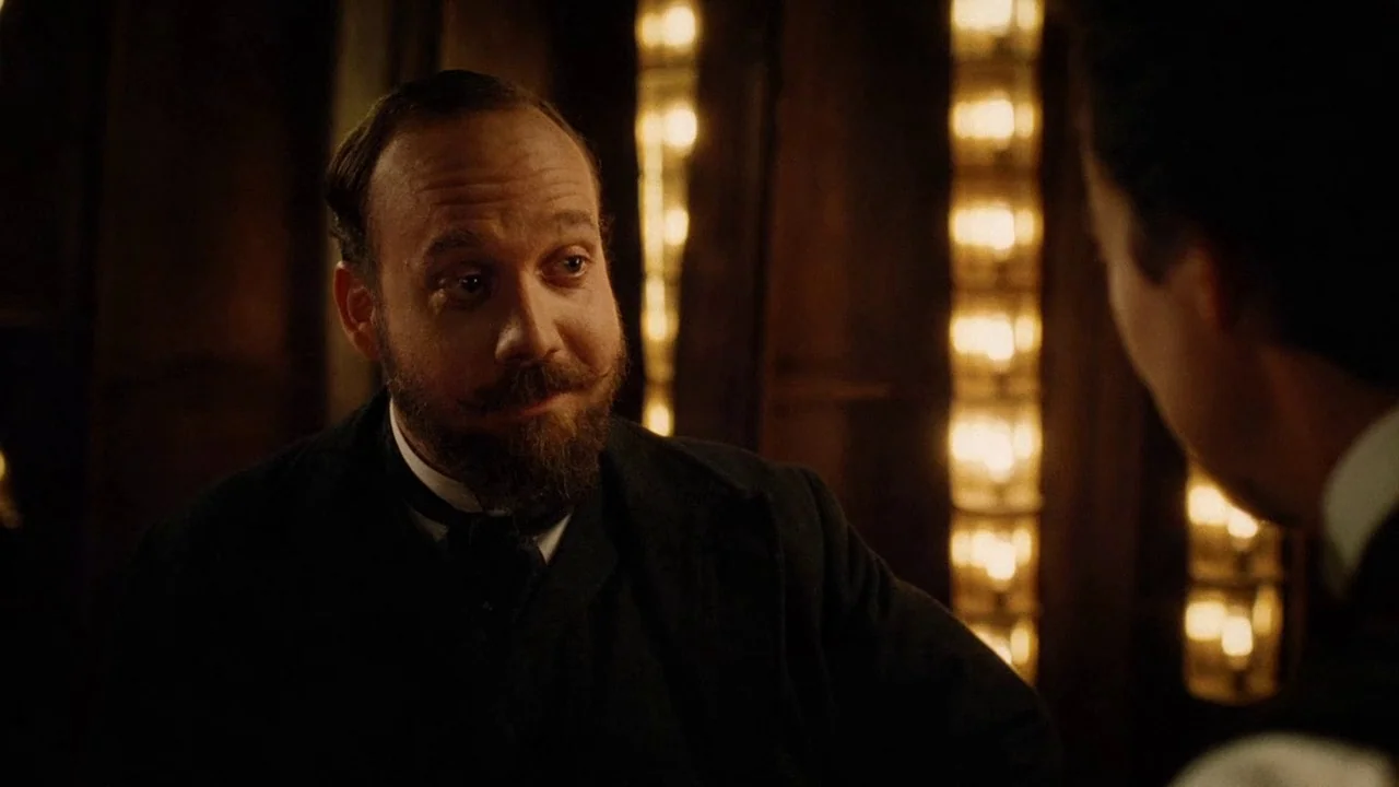 The Illusionist (2006) movie still: medium — A dimly lit room features a man with a mustache sitting near illuminated vanity bulbs. He sits in a…
