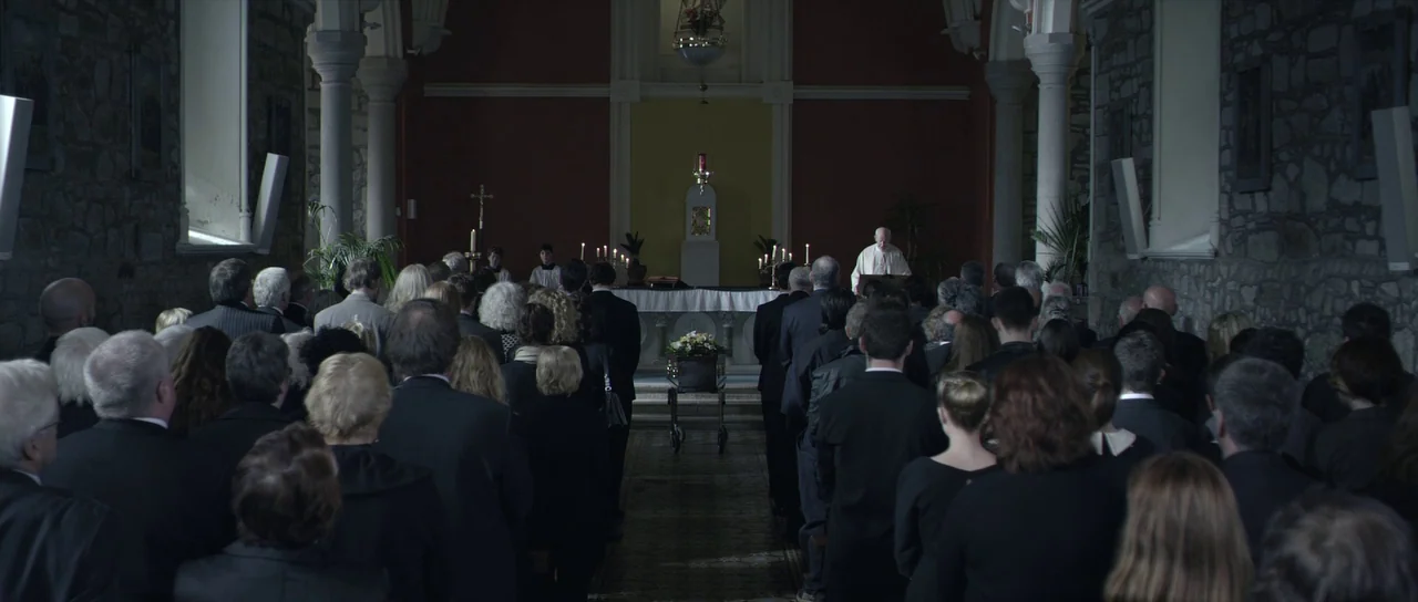 What Richard Did (2012) movie still: wide — A stone church nave with rows of people standing before a central altar. A wooden coffin sits on a…