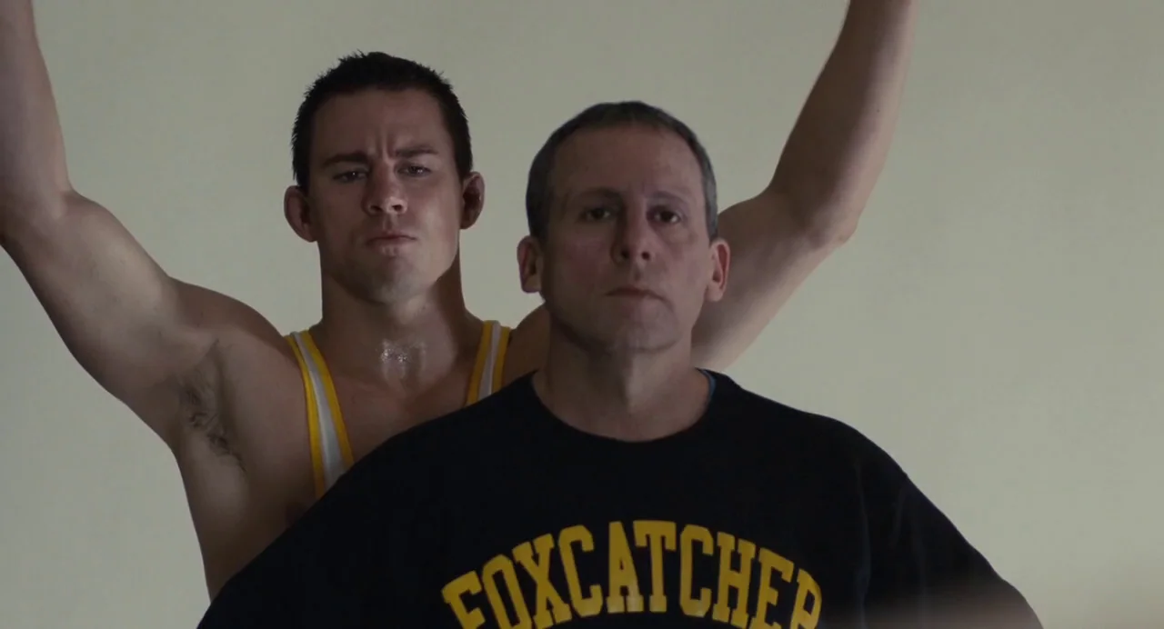 Foxcatcher (2014) movie still: medium — A man in a black shirt stands closely in front of a shirtless wrestler. The background is a plain off-white…