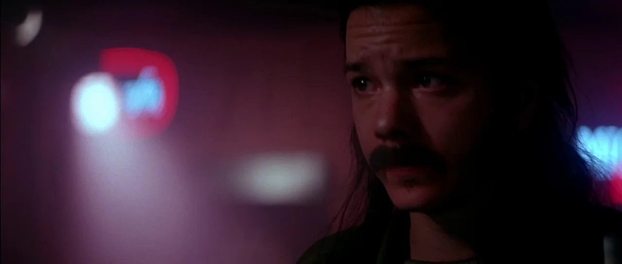 Born On The Fourth of July (1989) movie still: closeup — A dark, dimly lit bar environment with indistinct neon signage in the blurred background. A…
