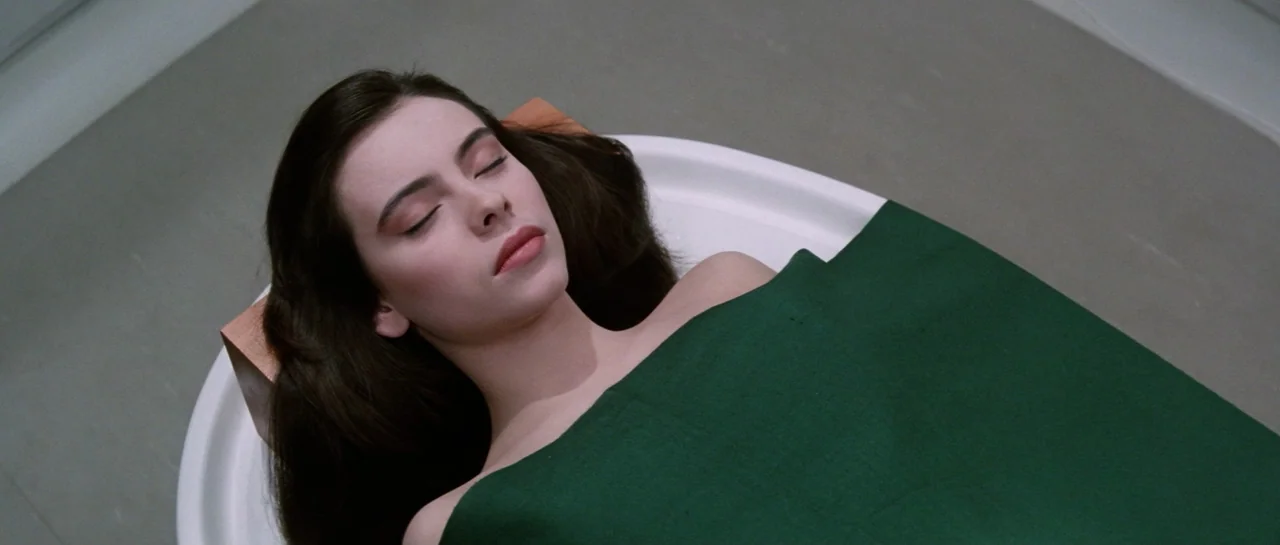 Lifeforce (1985) movie still: closeup — A woman lies on a white medical slab covered by a dark green fabric. The clinical space features neutral…