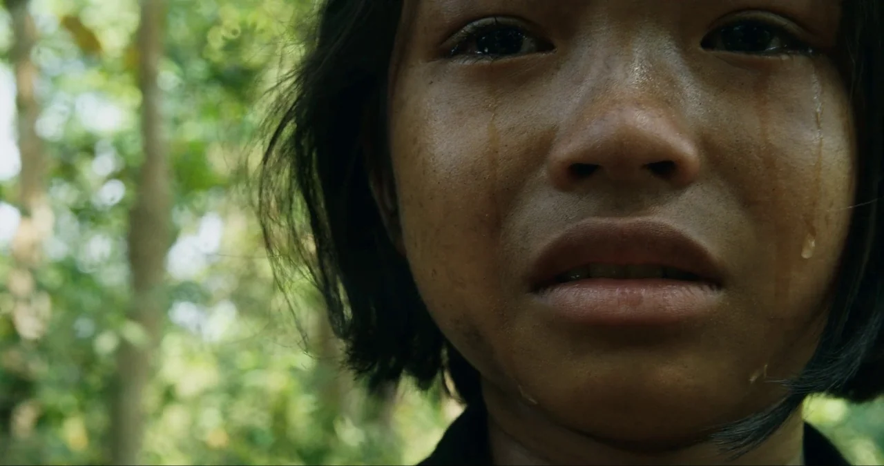 First They Killed My Father (2017) movie still: closeup — A dense green woodland provides a blurry backdrop for a close portrait. A young girl with…