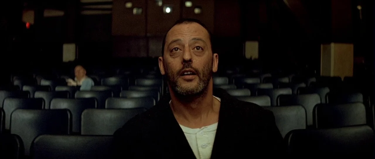 Léon: The Professional movie still: medium — Rows of empty dark seats fill a dimly lit movie auditorium. A middle-aged man sits in the foreground,…