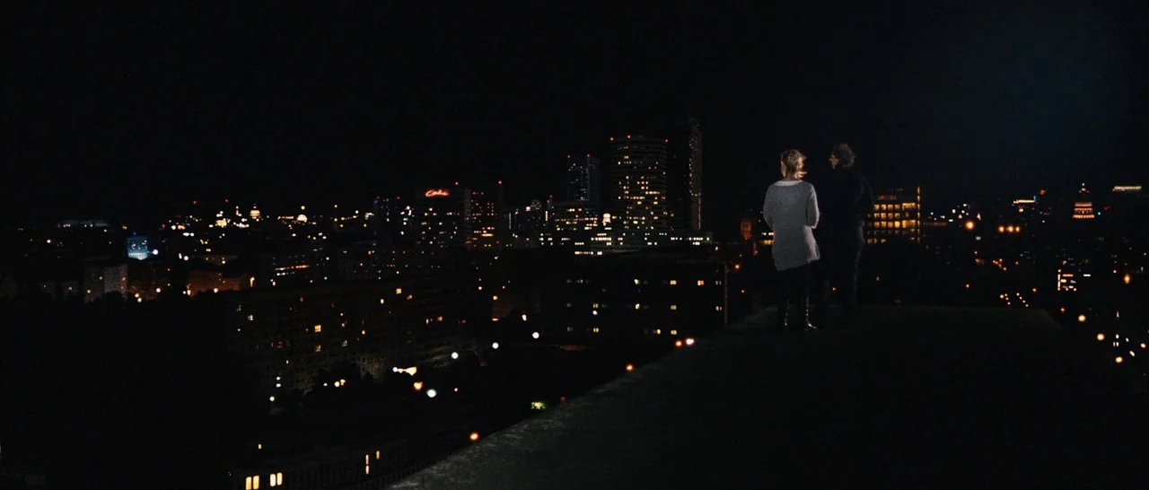 Sound of Noise (2010) movie still: wide — A flat concrete rooftop overlooks a sprawling city at night with scattered office lights. A man and a…