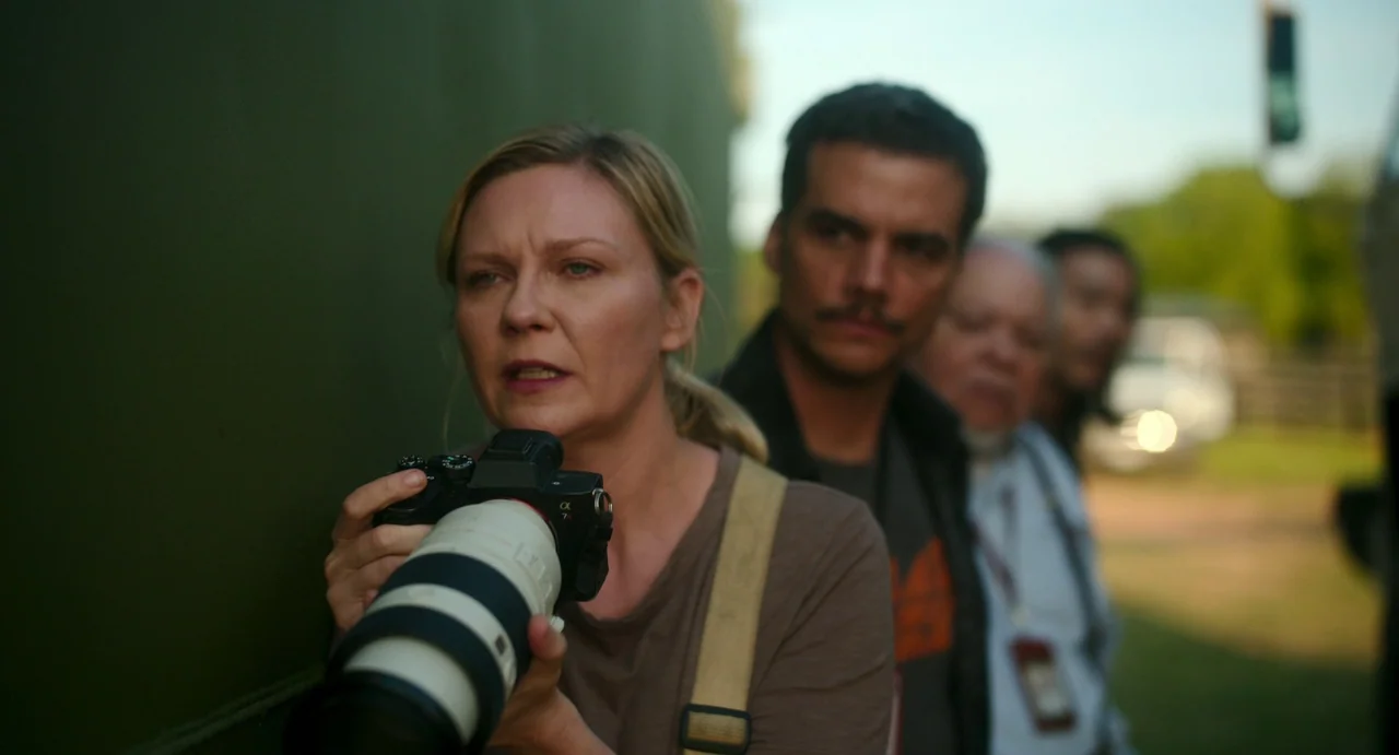 Civil War (2024) movie still: medium — A group of people stand in a line outdoors near a large olive-colored vehicle. A woman in the foreground aims…