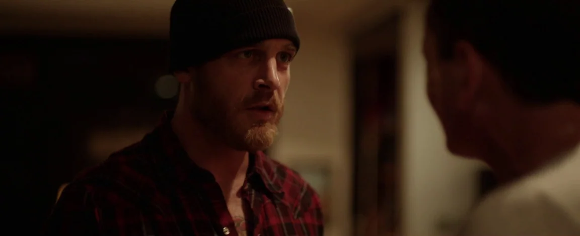 Cheap Thrills (2013) movie still: medium — A dimly lit room serves as the setting for an interaction between two men. The man in the foreground…