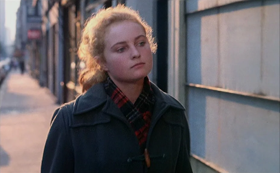 L’une Chante, l’Autre Pas (1977) movie still: medium — A woman walks along a paved sidewalk bordered by a plain building wall. Golden hour light…