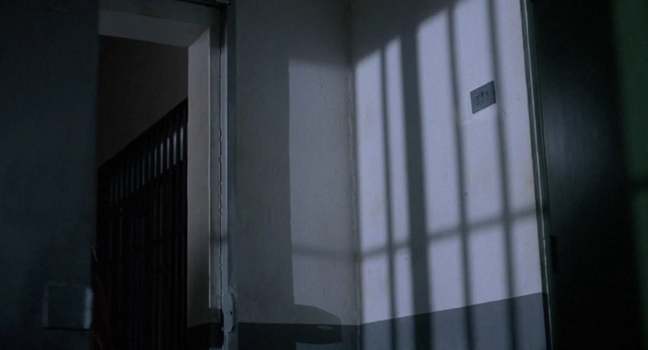 American Ninja (1985) movie still: establishing — A stark prison cell interior features plain white walls and a dark grey baseboard. Shadows from…