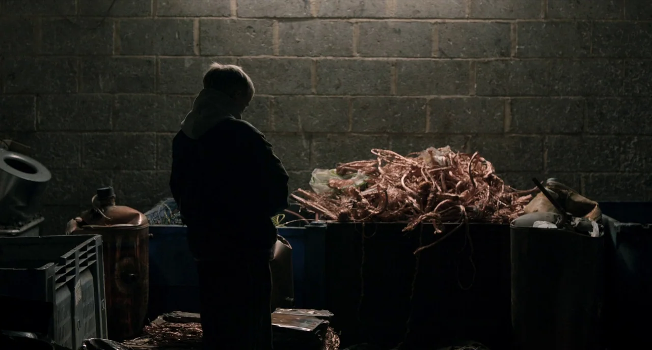The Selfish Giant (2013) movie still: fullbody — A dark indoor space with gray concrete block walls contains several large industrial containers. A…