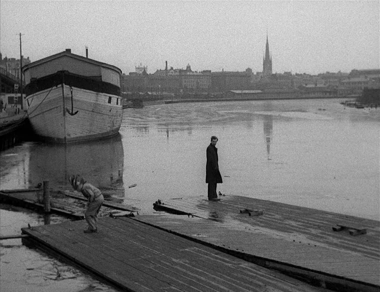 Prison (1949) movie still: wide — An industrial waterfront features floating wooden piers on a murky, overcast river. A man stands on the pier as a…