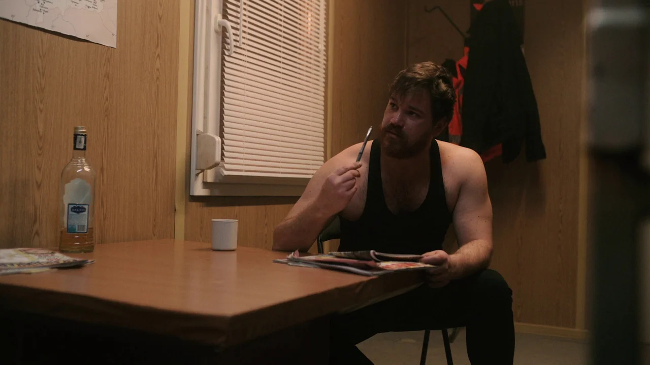 Helll Hole (2024) movie still: medium — A small room features paneled walls and a table with a bottle and magazines. A man in a black tank top sits…