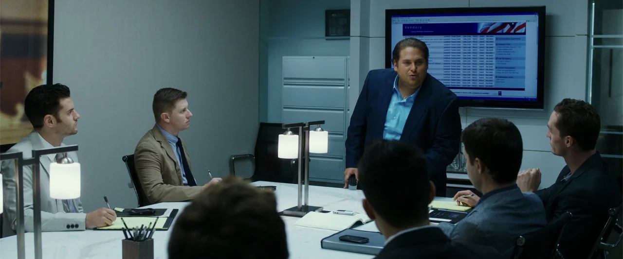 War Dogs (2016) movie still: medium — A modern corporate workspace with a white marble table and a wall-mounted flat-screen display. A man in a blue…