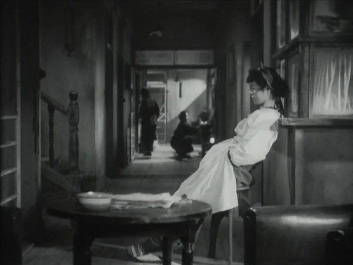 The Quiet Duel (1949) movie still: fullbody — A dim, narrow hallway with wooden floorboards and a central stairwell. A woman in a light dress stands…