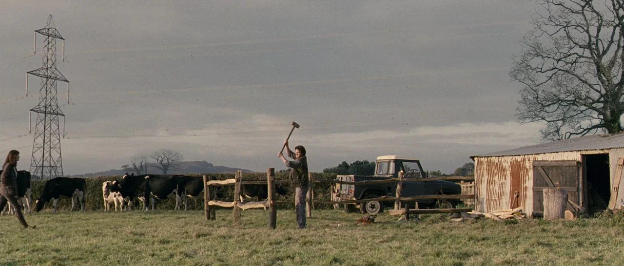 Tamara Drewe (2010) movie still: wide — A rural pasture contains a wooden fence, cattle, a utility truck, and a shed. A man swings a sledgehammer at…