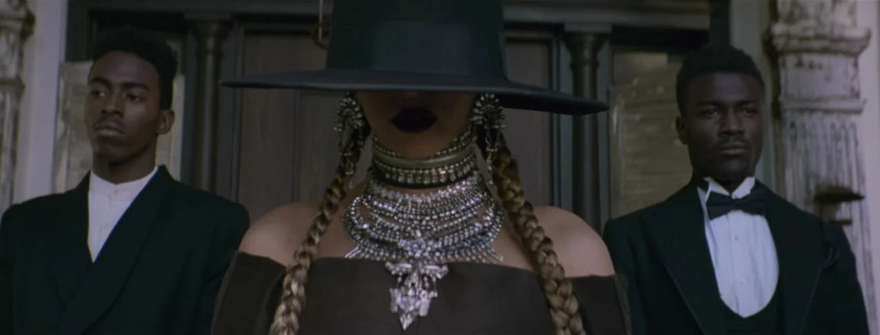 Lemonade (2016) movie still: medium — A woman stands centered between two men in front of a wood-paneled entrance. The subjects wear formal attire…