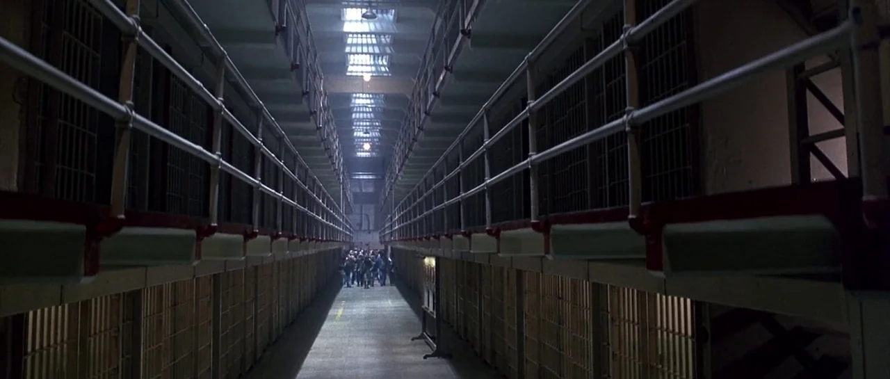 The Rock (1996) movie still: wide — A long, symmetrical prison block with multiple levels of steel barred cells. A group of men stands at the far…