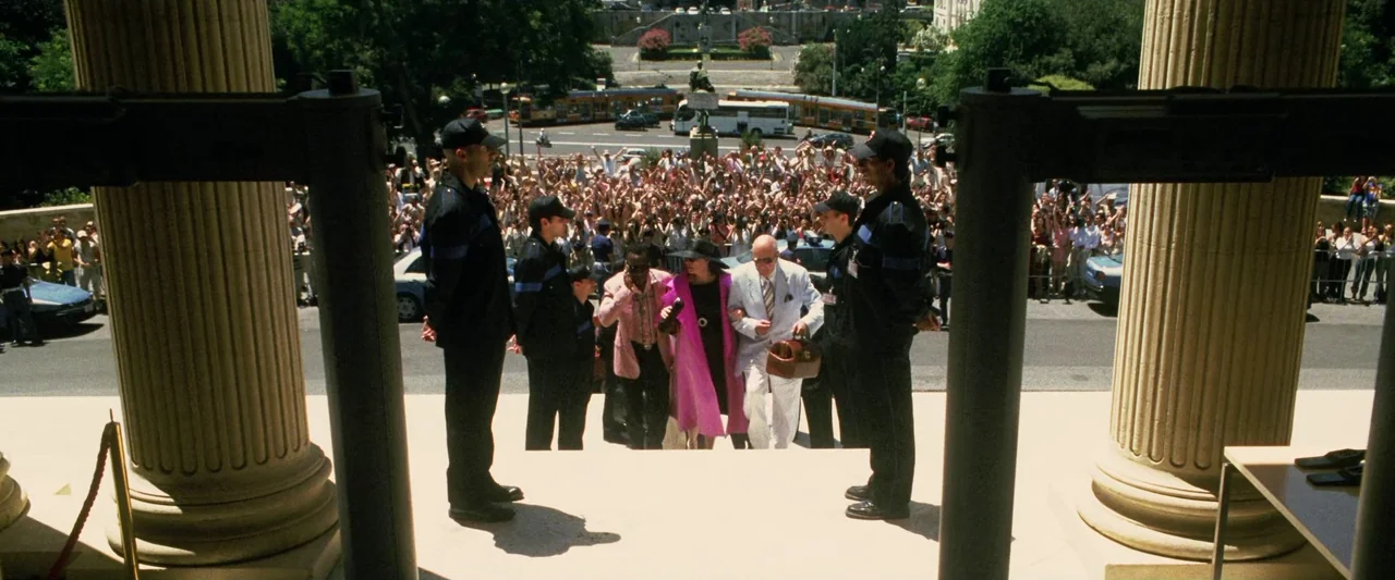 Ocean’s Twelve (2004) movie still: wide — A stone building entrance with large columns and metal security scanners stands before a crowd. A woman in…