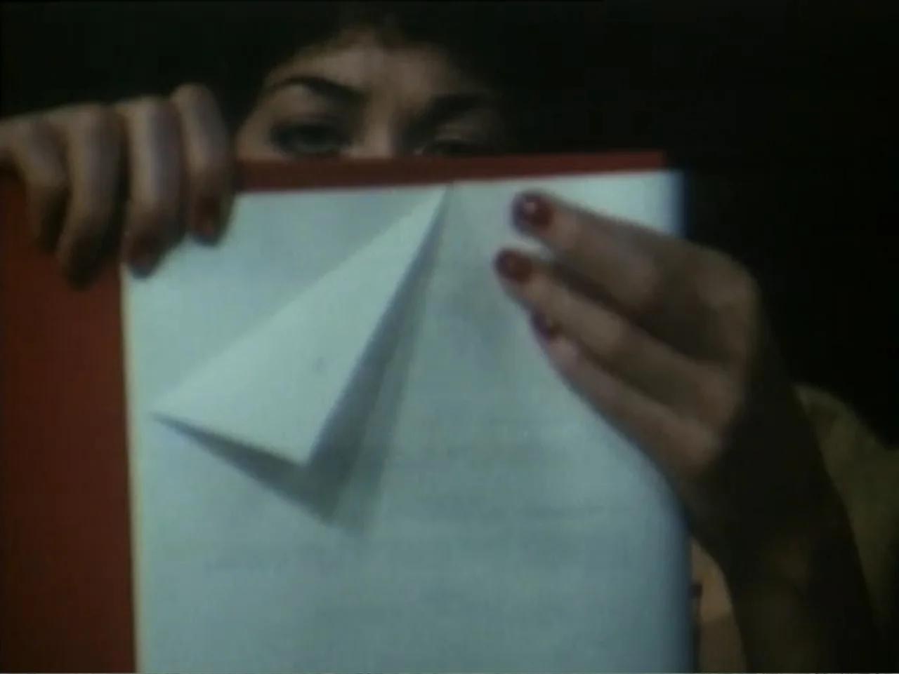 Curtains (1995) movie still: closeup — A dark indoor space contains a person holding a red folder. The subject peers over the top of the paper to…