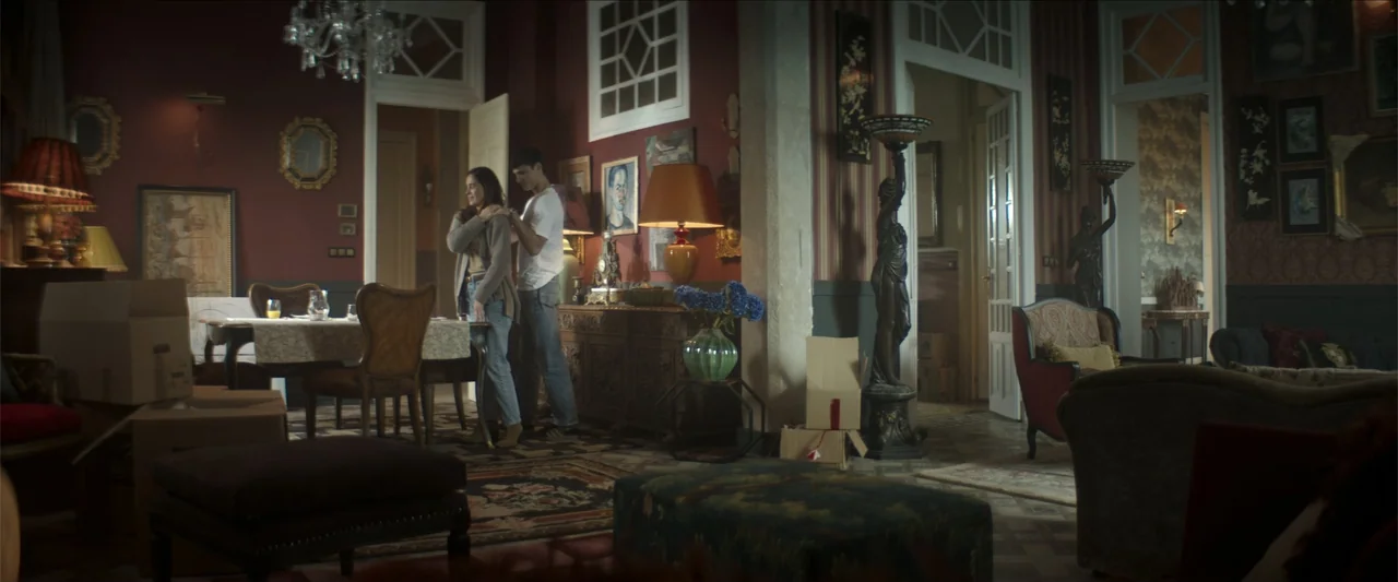 Tell Me Softly (2025) movie still: fullbody — A spacious room features dark red walls, ornate antique furniture, and unpacked cardboard boxes. A man…