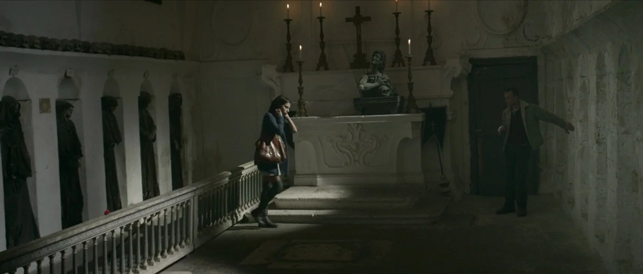 Spring (2014) movie still: wide — A stone subterranean room features wall niches containing robed figures and a central altar. A woman stands near a…